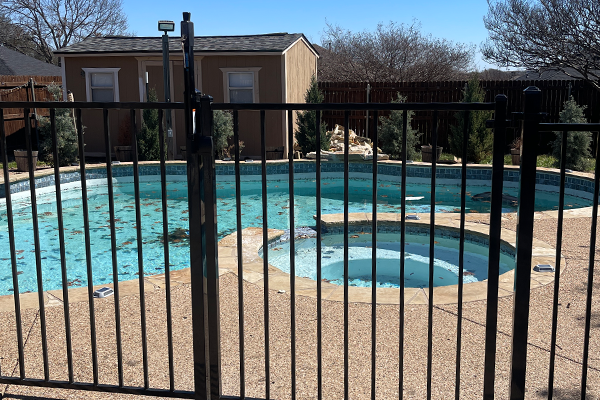 Black fenced-in, dirty pool; small building in the background. Clear sky.
