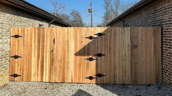 Wooden gate with black hinges, between brick walls, gravel ground.