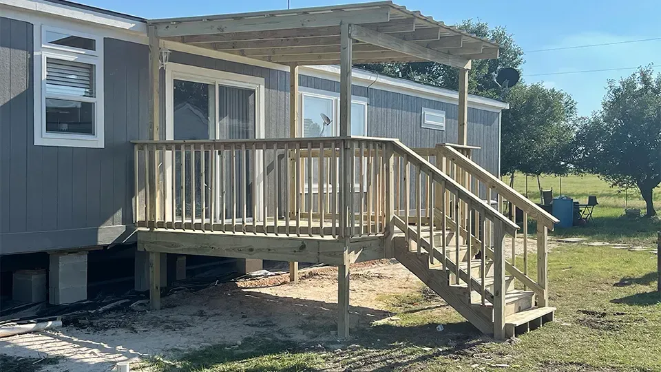 Wooden deck with stairs and pergola attached to a gray house with a window and sliding glass door.