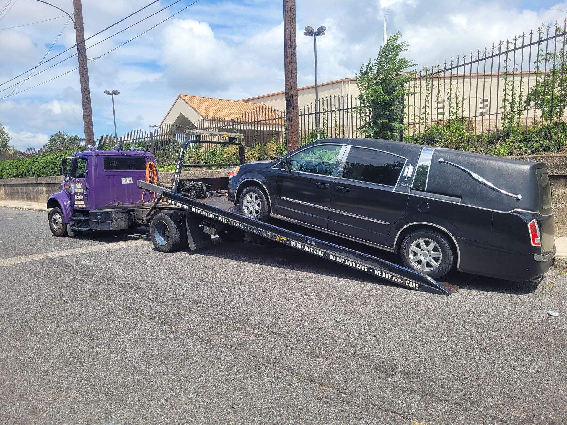 Black hearse on a tow truck with a purple towing vehicle on a street near a fence