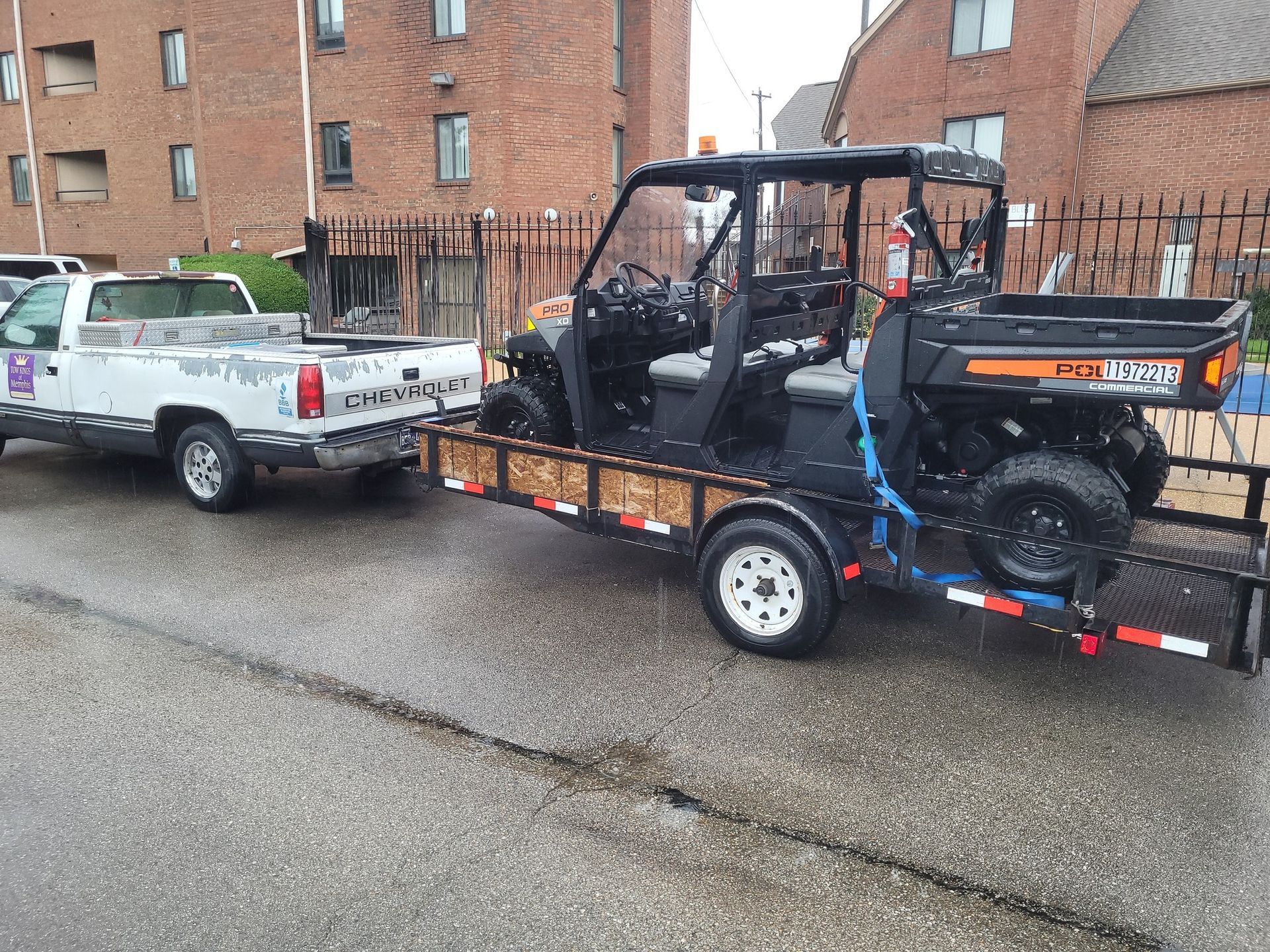 White pickup truck towing a black ATV trailer in a wet parking lot beside brick apartments