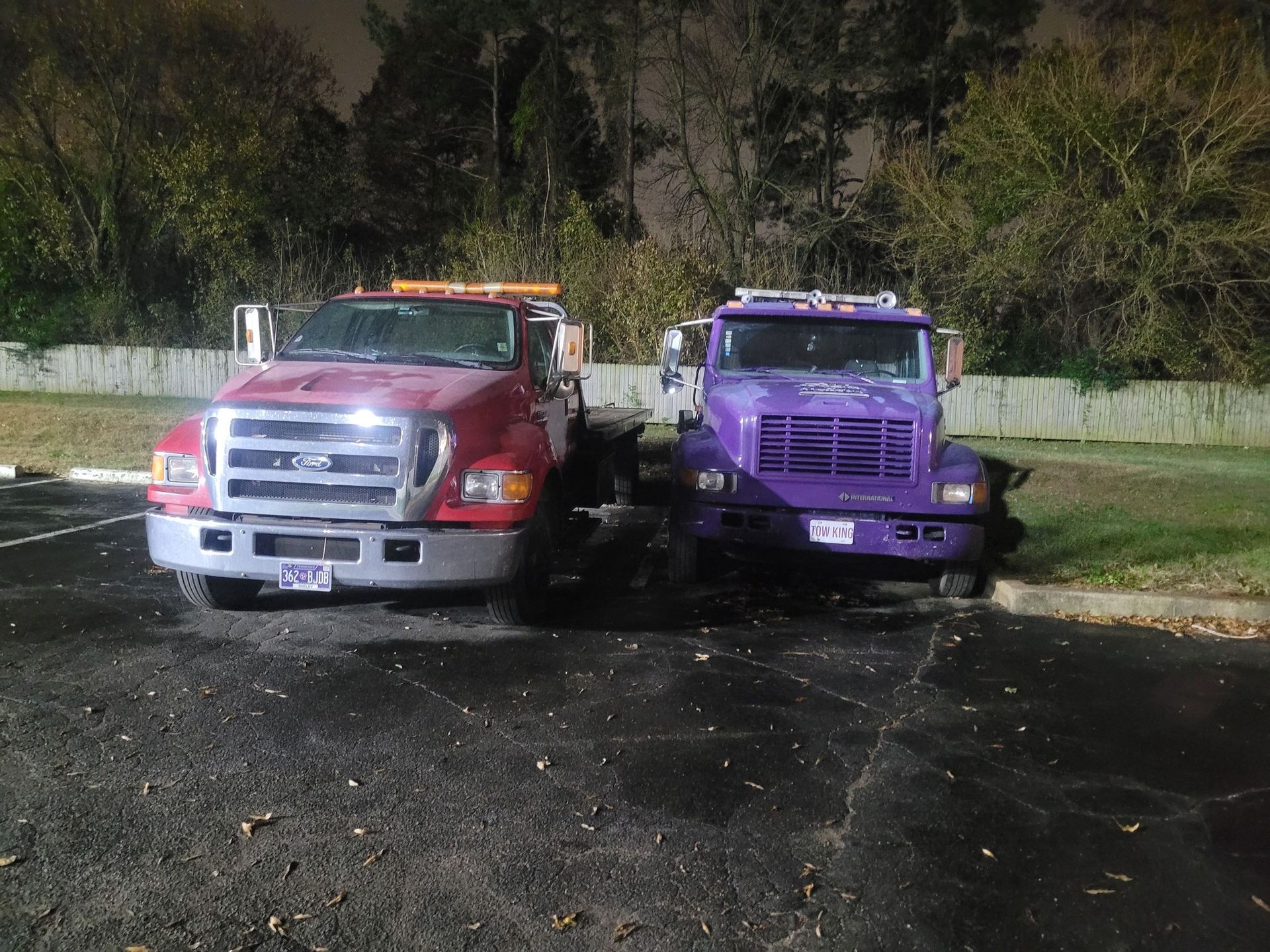 Two parked tow trucks, one red and silver and one purple, at night on a wet lot