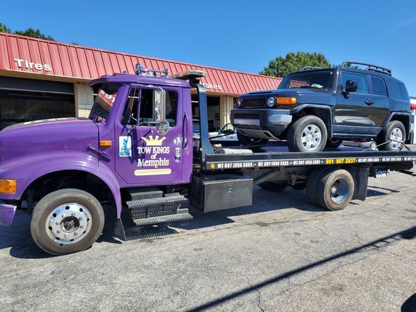 Purple tow truck carrying a black SUV outside a building with a red roof