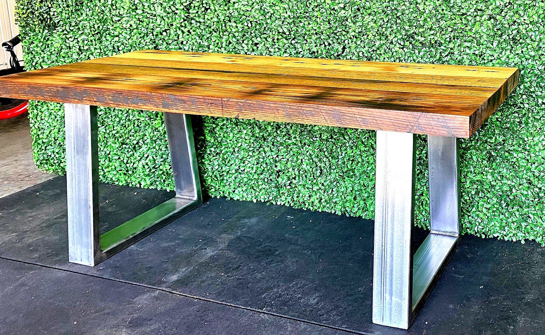 A wooden table with metal legs is sitting on a black mat in front of a hedge.