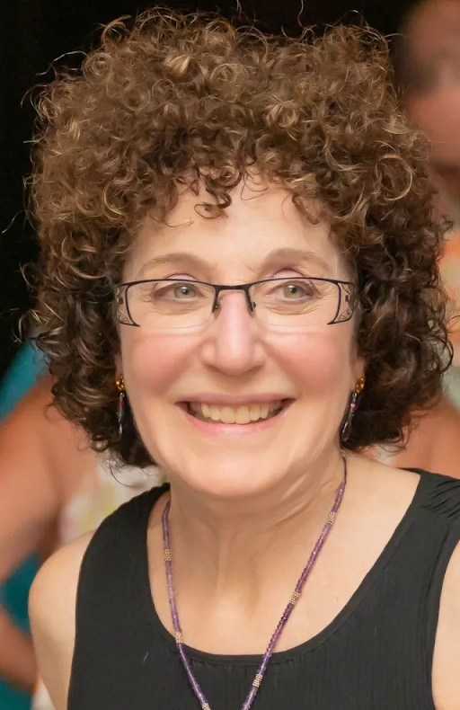 Woman with curly brown hair and glasses smiles, wearing a black top and purple necklace.