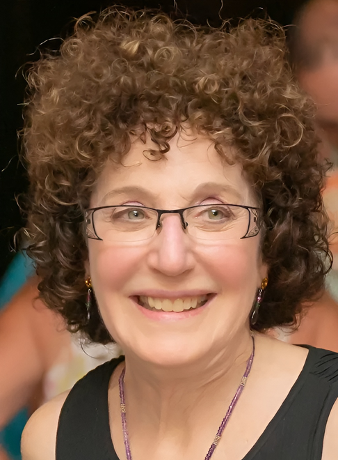 Woman with curly brown hair and glasses smiles, wearing a black top and purple necklace.