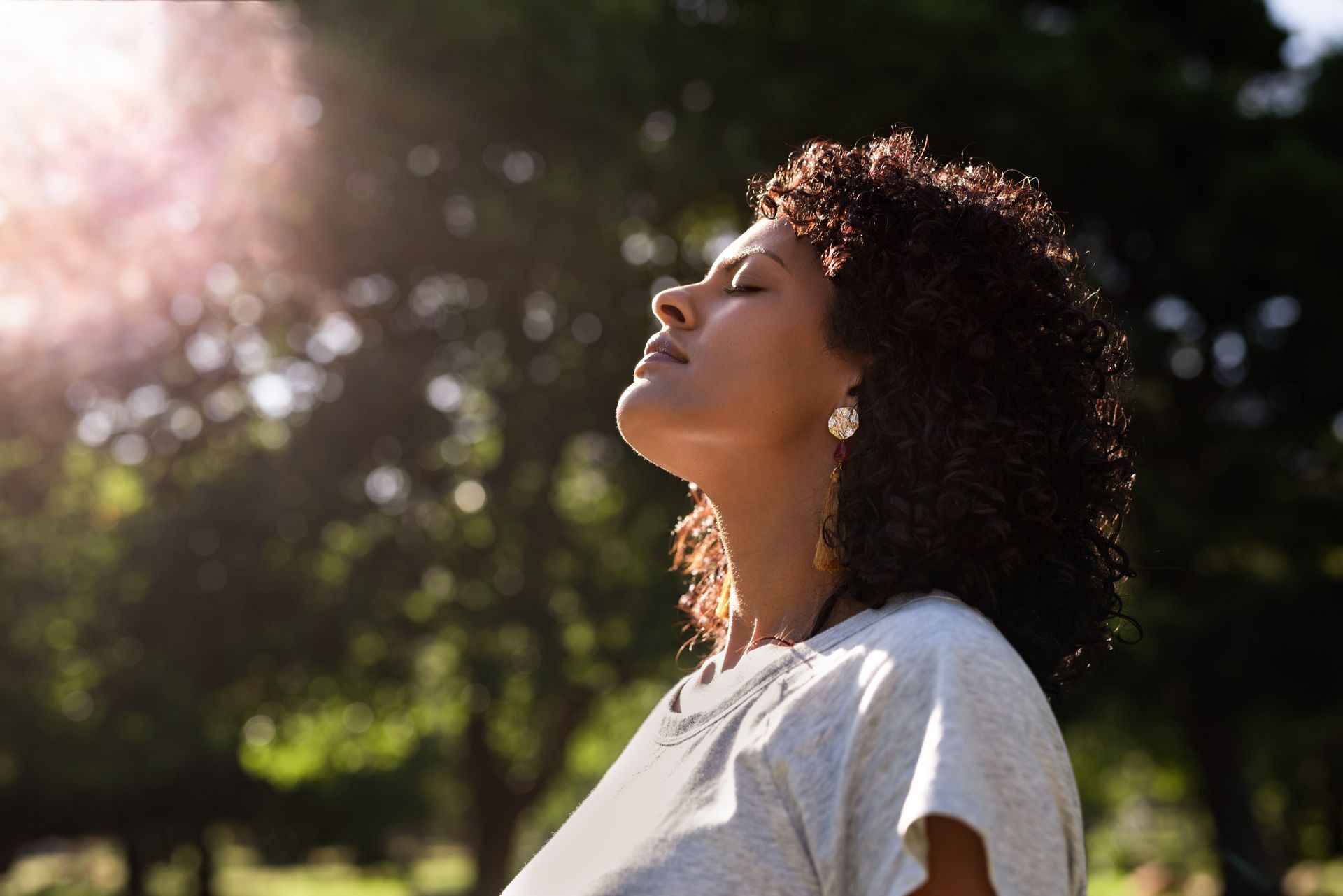 Woman outdoors with eyes closed, facing sunlight among blurred trees