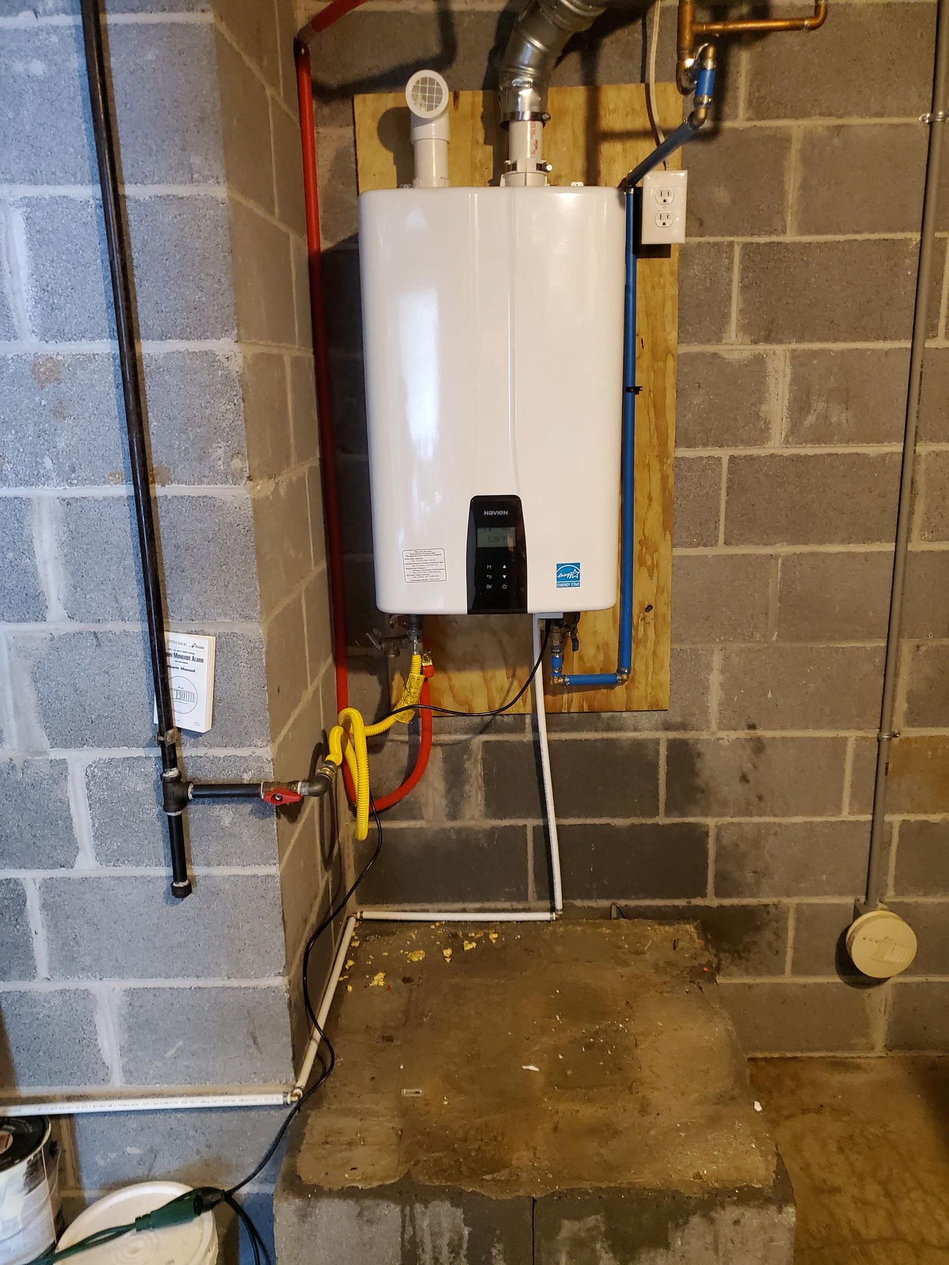 A white water heater is mounted to a brick wall in a basement.