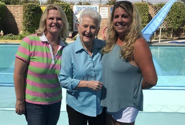 Three people standing side by side smiling, with an outdoor swimming pool and slide in the background.