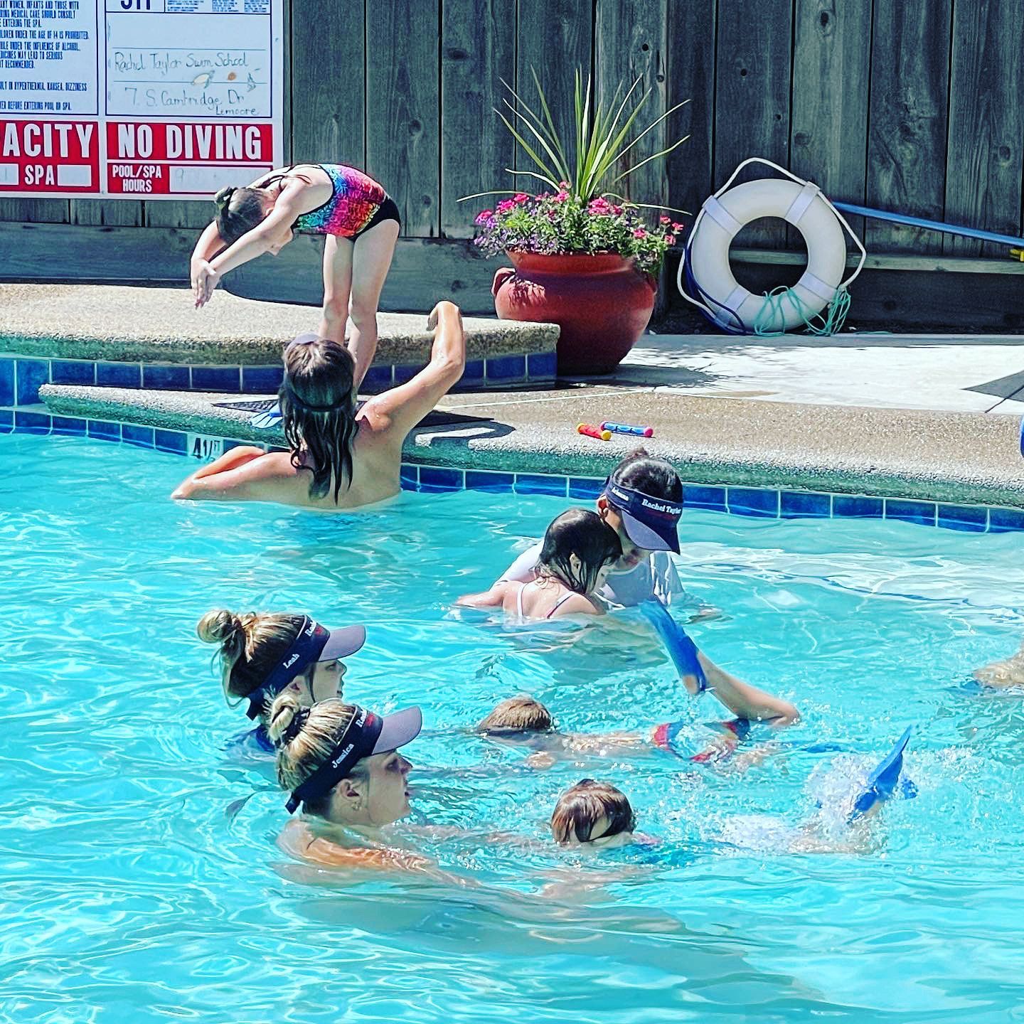 A group of people in a pool, with some learning to swim using blue kickboards, while a child prepares to dive nearby.