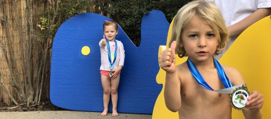 Two children wearing medals and giving thumbs-up gestures in front of large blue and yellow shapes.