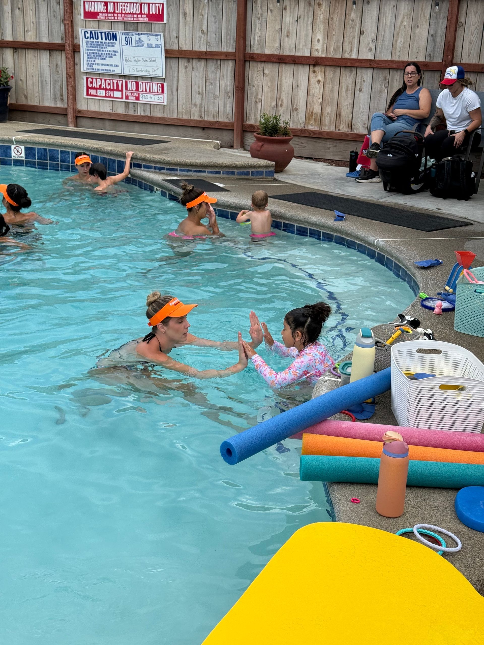 An instructor wearing an orange visor guides a child in a patterned swimsuit through water in a backyard swimming pool.