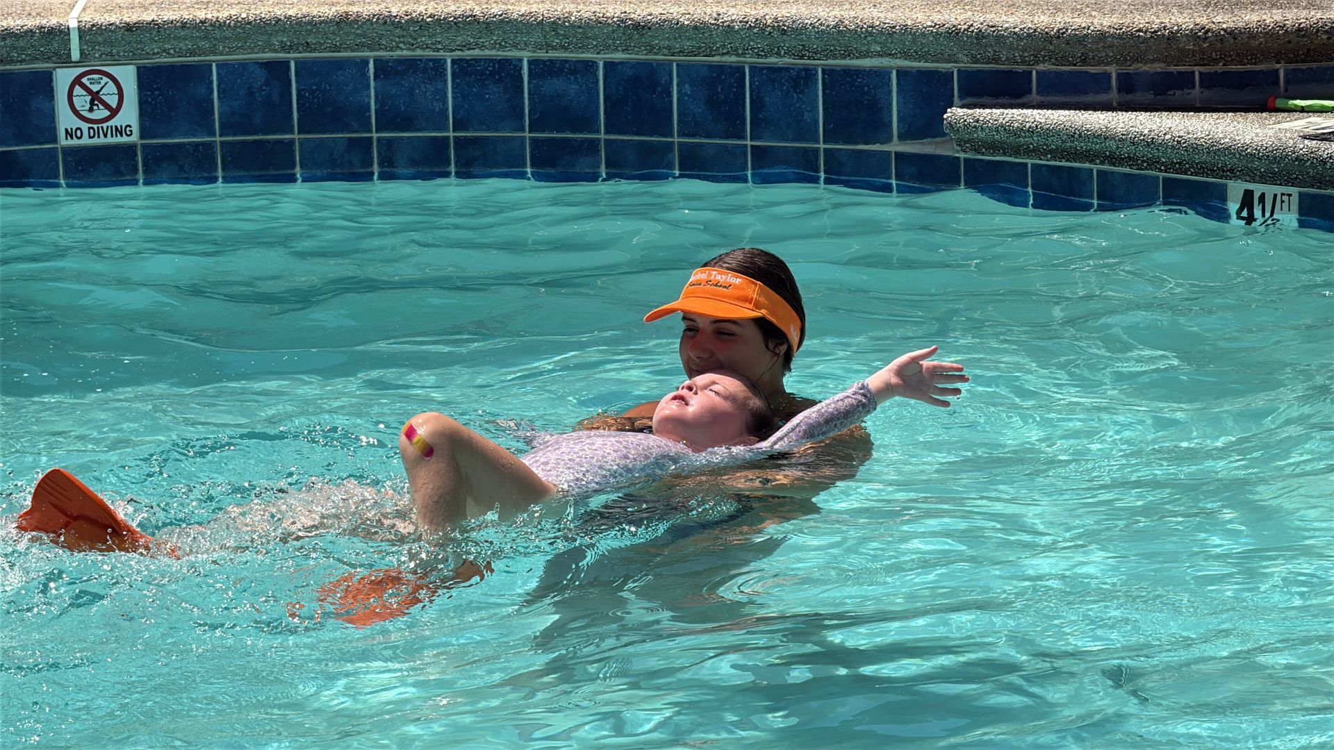 An adult helps a child practice floating in a swimming pool, with both wearing orange accessories.