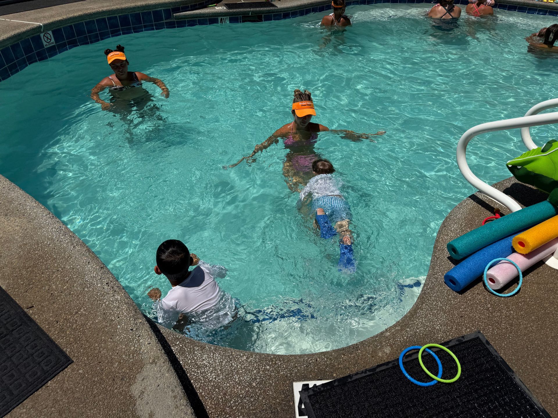 People wearing orange visors swim and play in a backyard pool with colorful foam noodles and diving rings nearby.