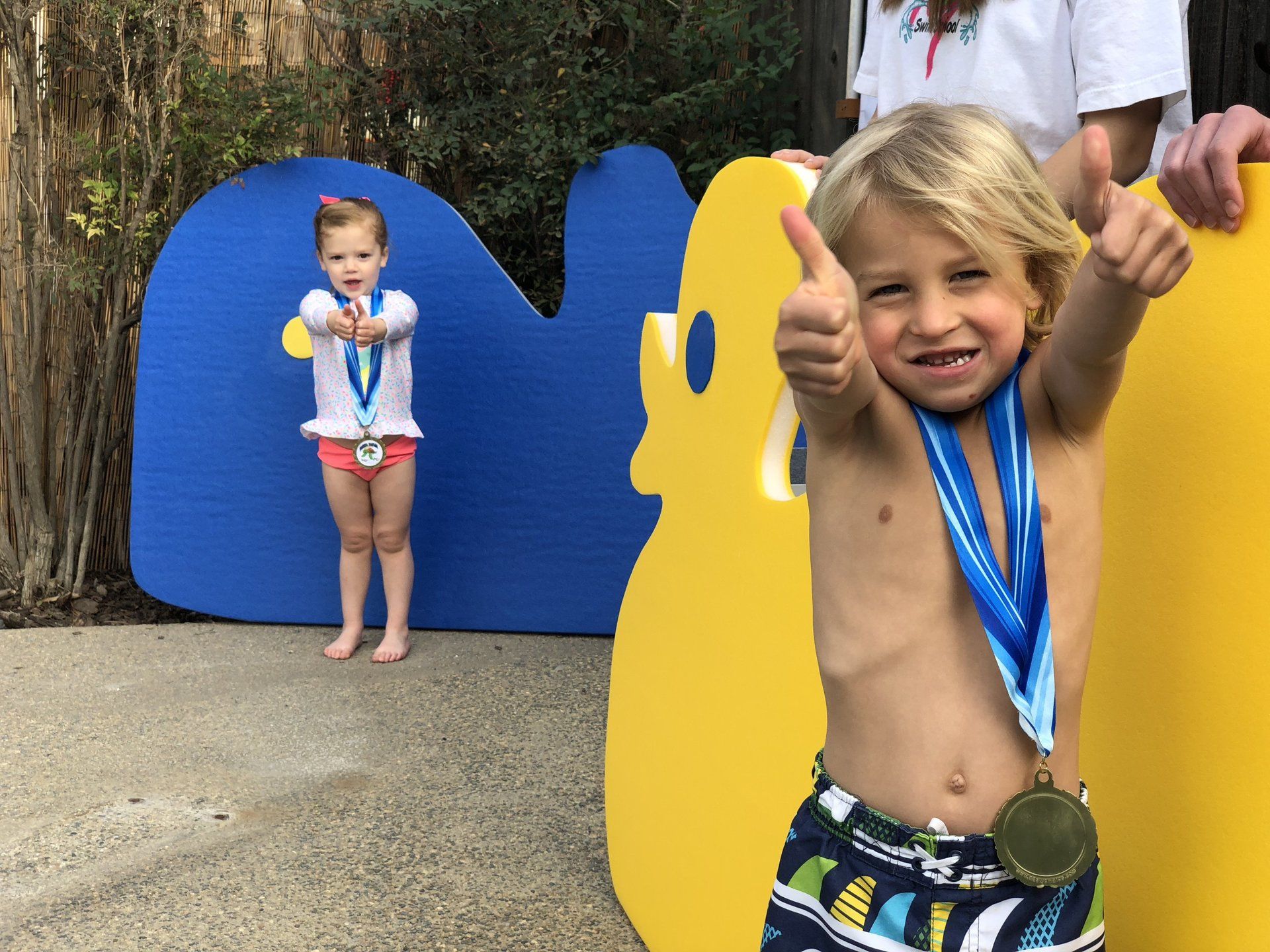 Two children wearing medals and giving thumbs up in front of large blue and yellow cutouts.
