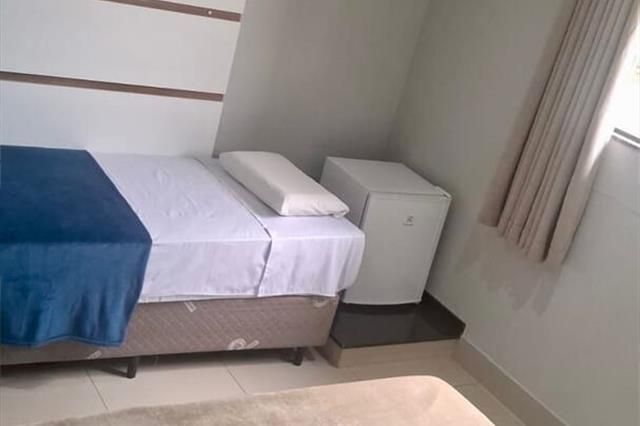 Quarto pequeno com cama, cobertor azul, mesa de cabeceira e cortinas bege junto à janela.