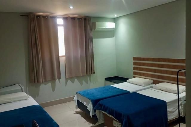 Quarto de hotel pequeno com três camas de solteiro, janela com cortina e ar condicionado em paredes verde-claras.