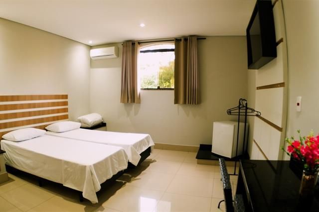Quarto de hotel minimalista com duas camas de solteiro, paredes bege, cortinas e um pequeno suporte para TV.
