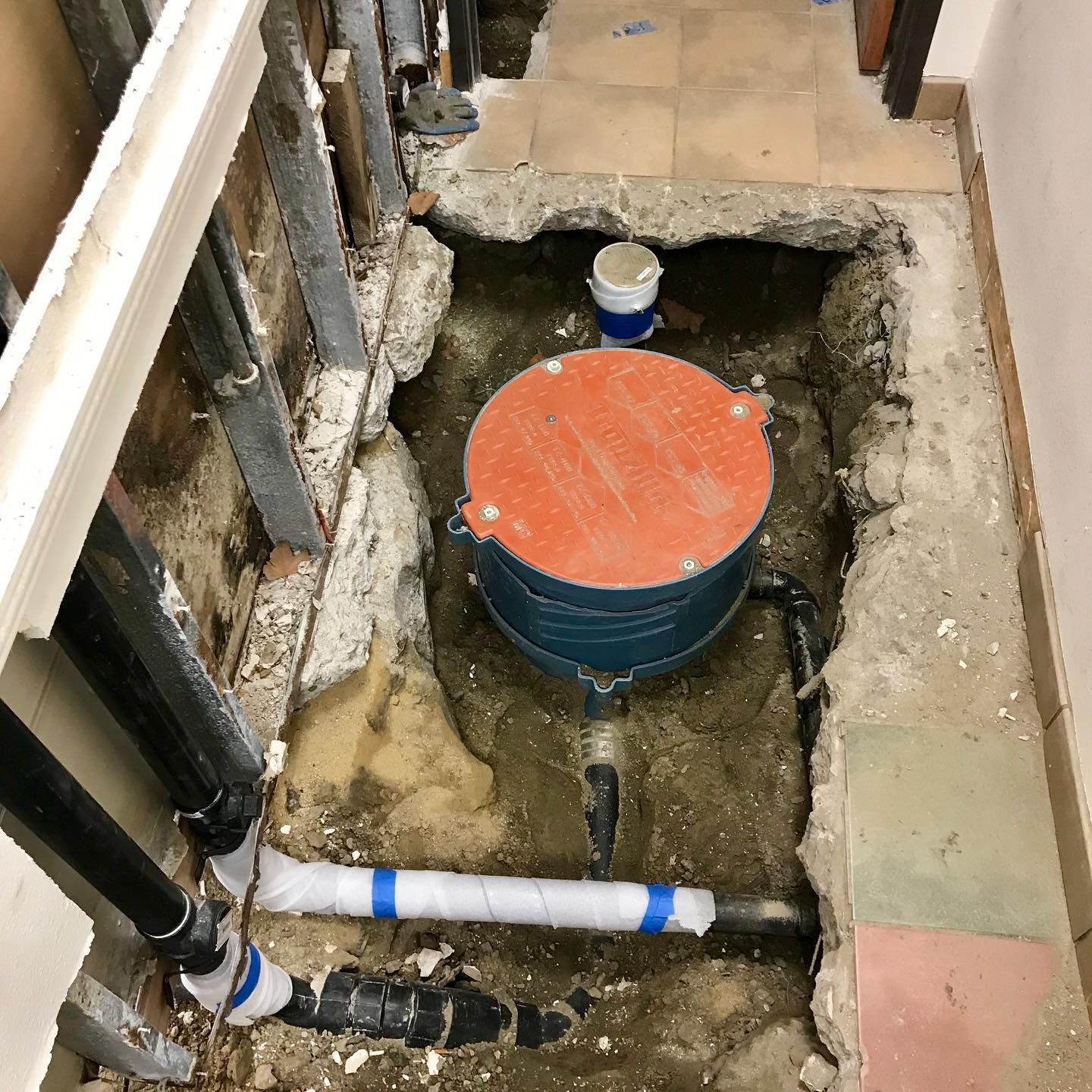 Excavation in a hallway, showing a blue water tank with an orange lid. Black and white pipes exposed. Concrete and drywall.