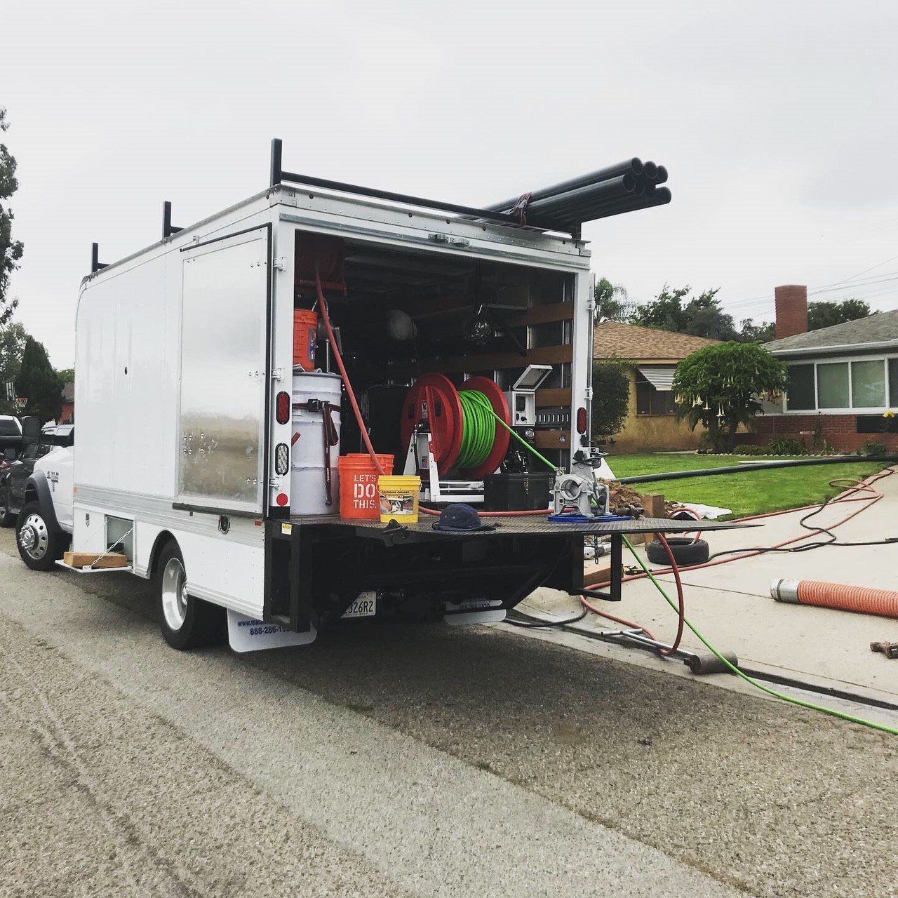 White utility truck with open back on a residential street, equipment visible inside and on the ground.