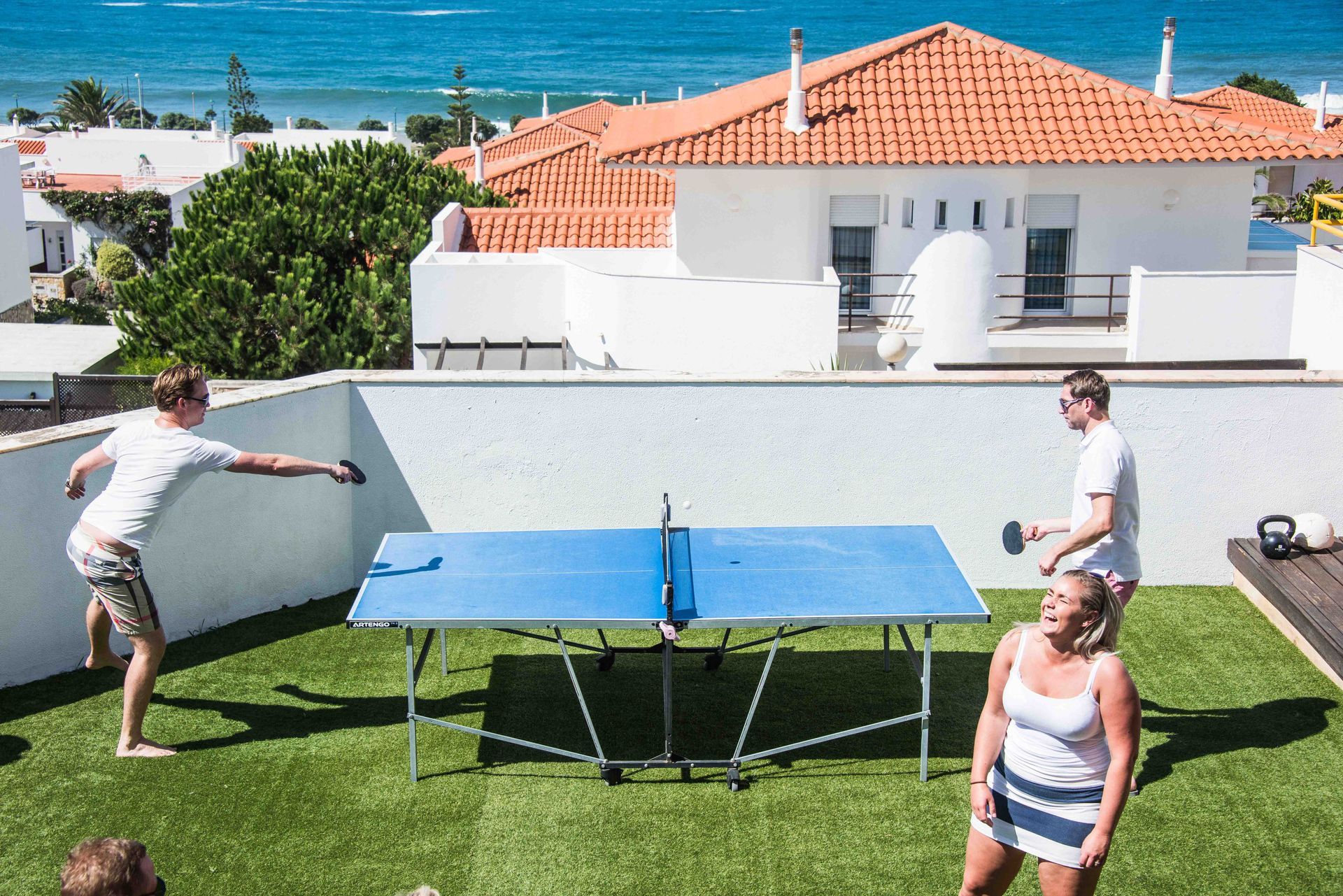 En gruppe mennesker spiller bordtennis på et hustak med utsikt over havet.