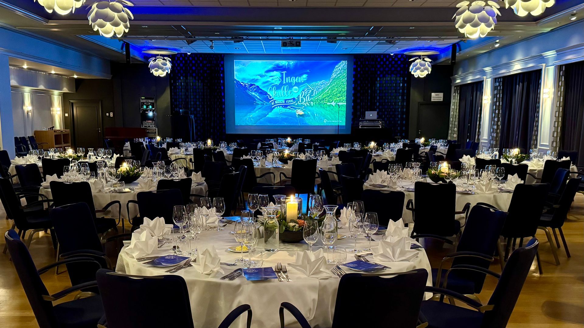 Et stort rom fylt med bord og stoler og en stor skjerm.