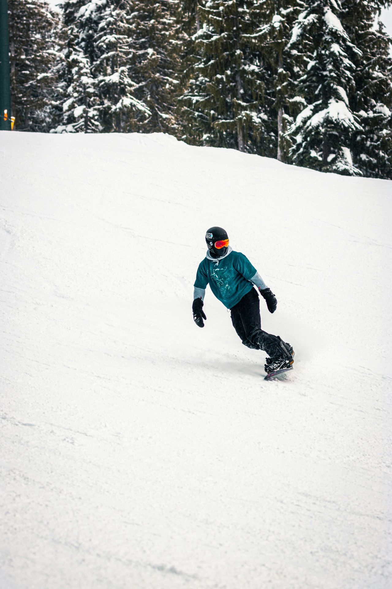 Person i neongrønne skibukser og grønn lue sitter på en snødekt bakke og ser ned; skiheis i bakgrunnen.