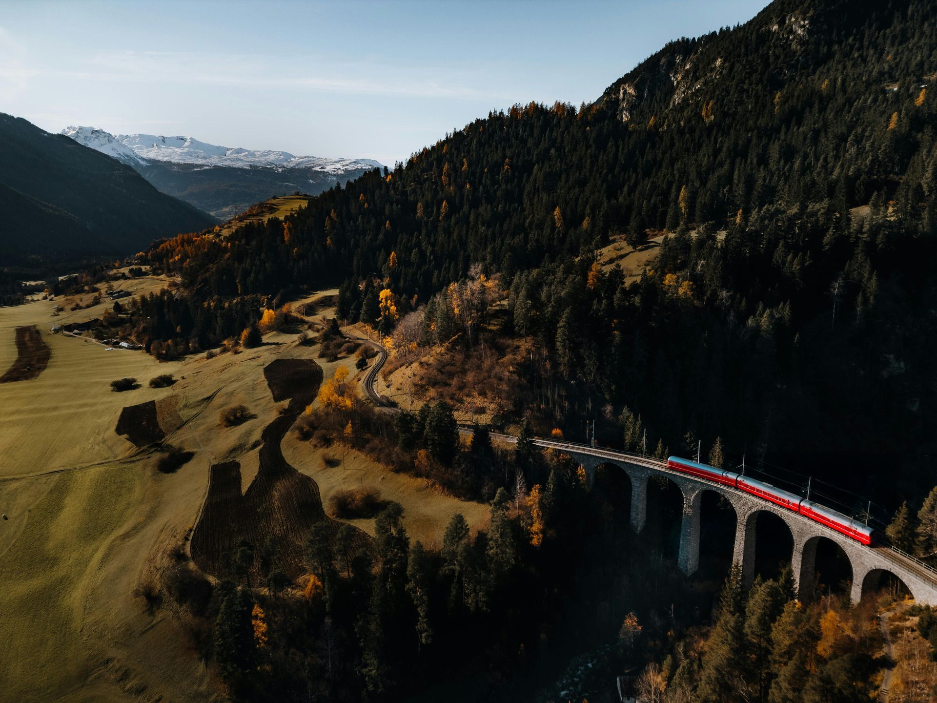Train traveling across a curved stone bridge in a valley, surrounded by mountains and trees; autumn colors.