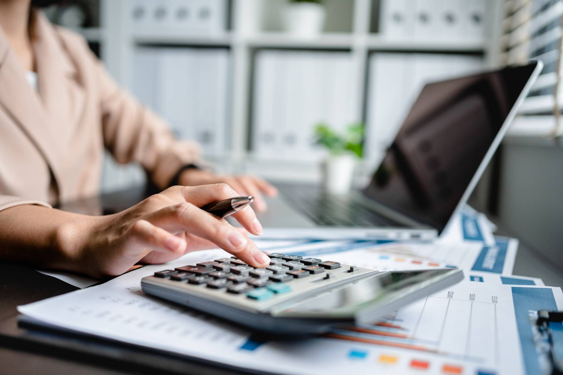 Hands using a calculator on financial charts beside an open laptop and pen