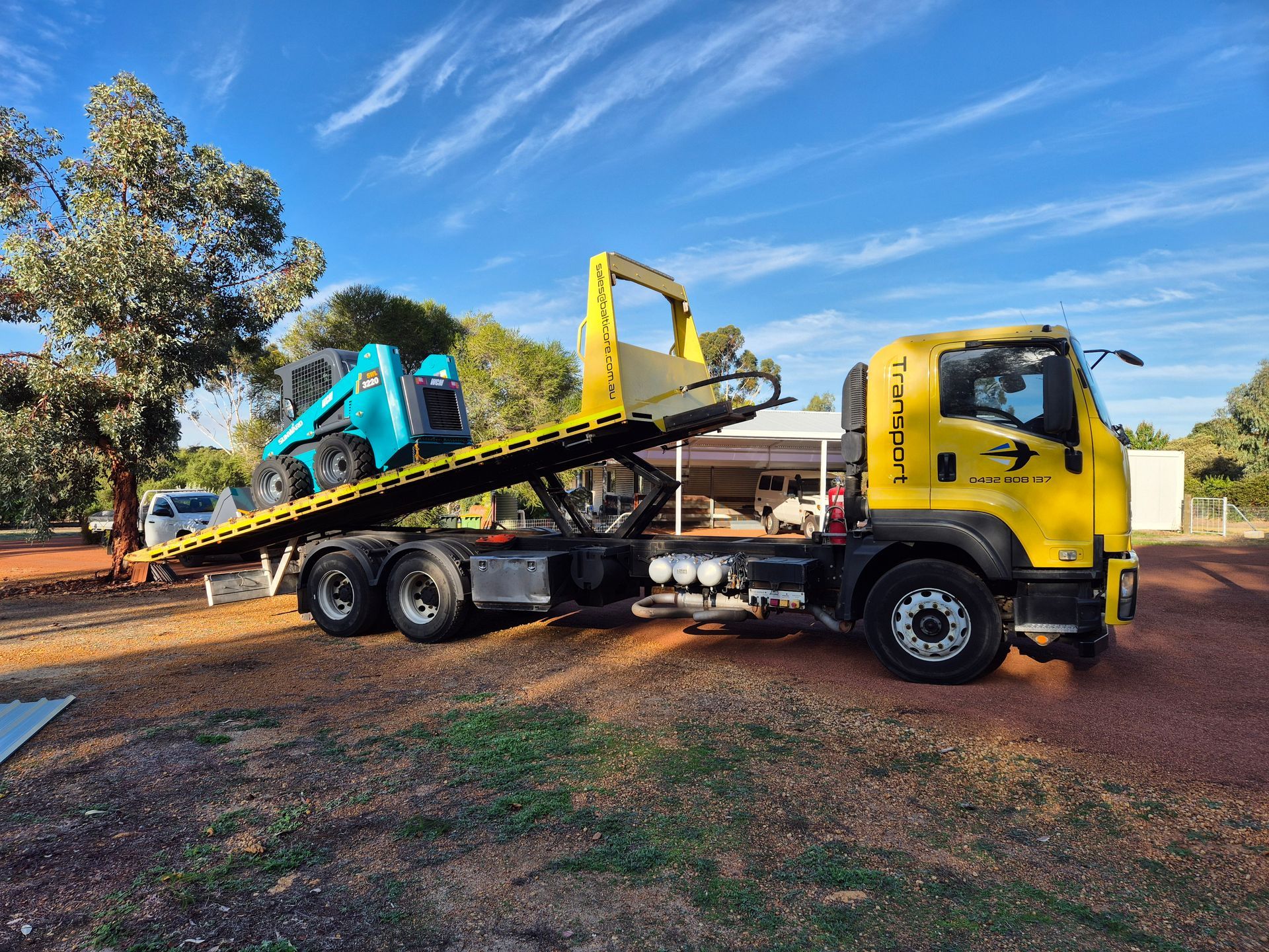 Yellow tow truck carrying a blue skid steer on its flatbed, parked on red dirt under a blue sky.