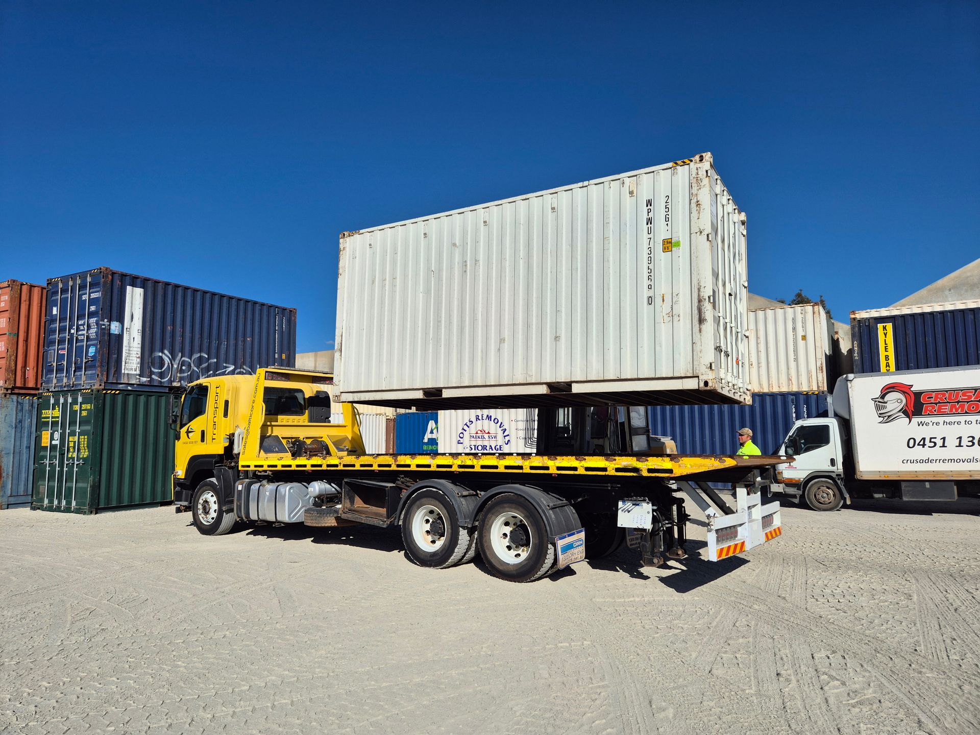 Yellow flatbed truck carrying a white shipping container in a yard with other containers under a blue sky.