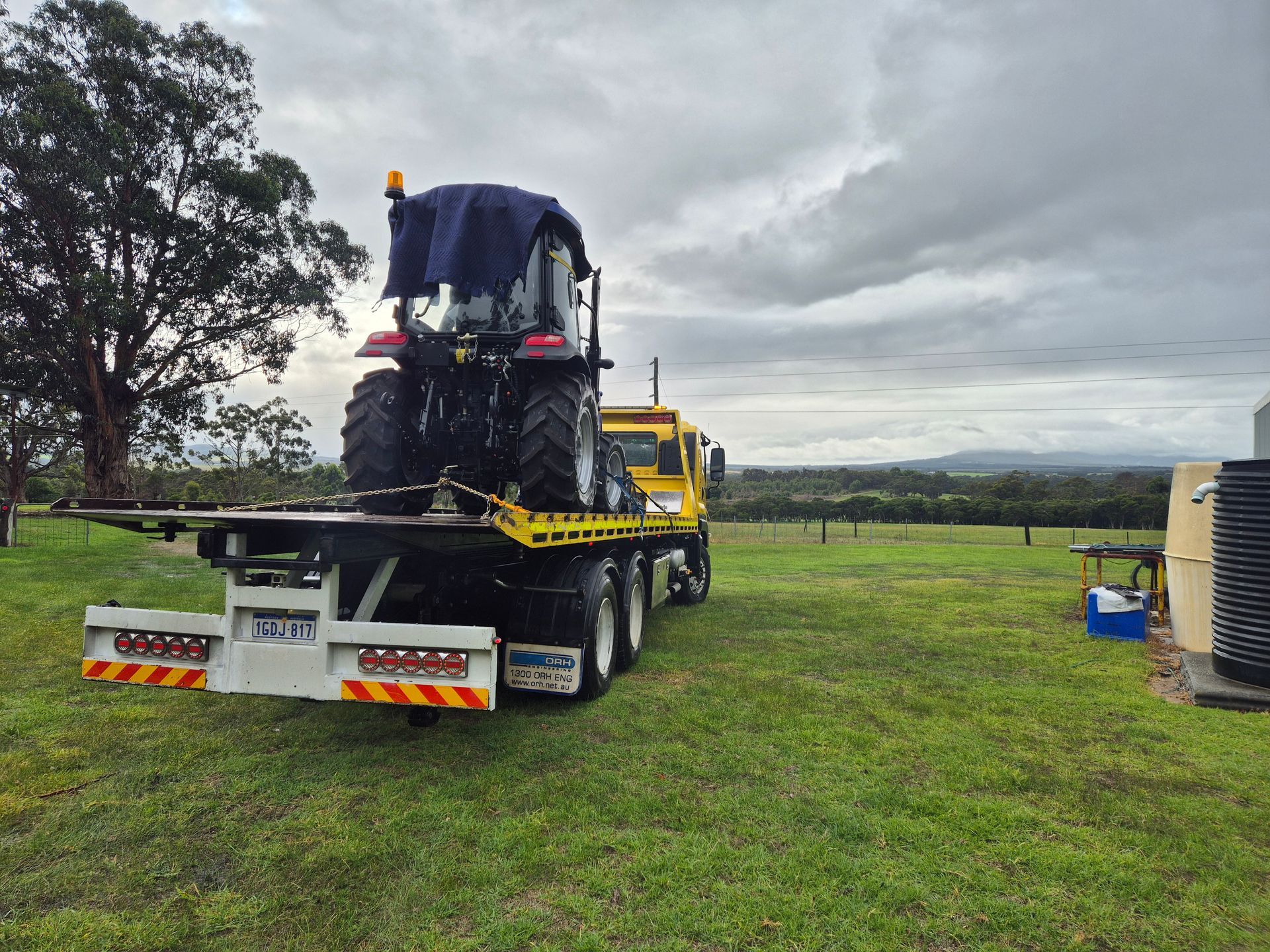 A tractor on a flatbed truck in a grassy field under a cloudy sky.