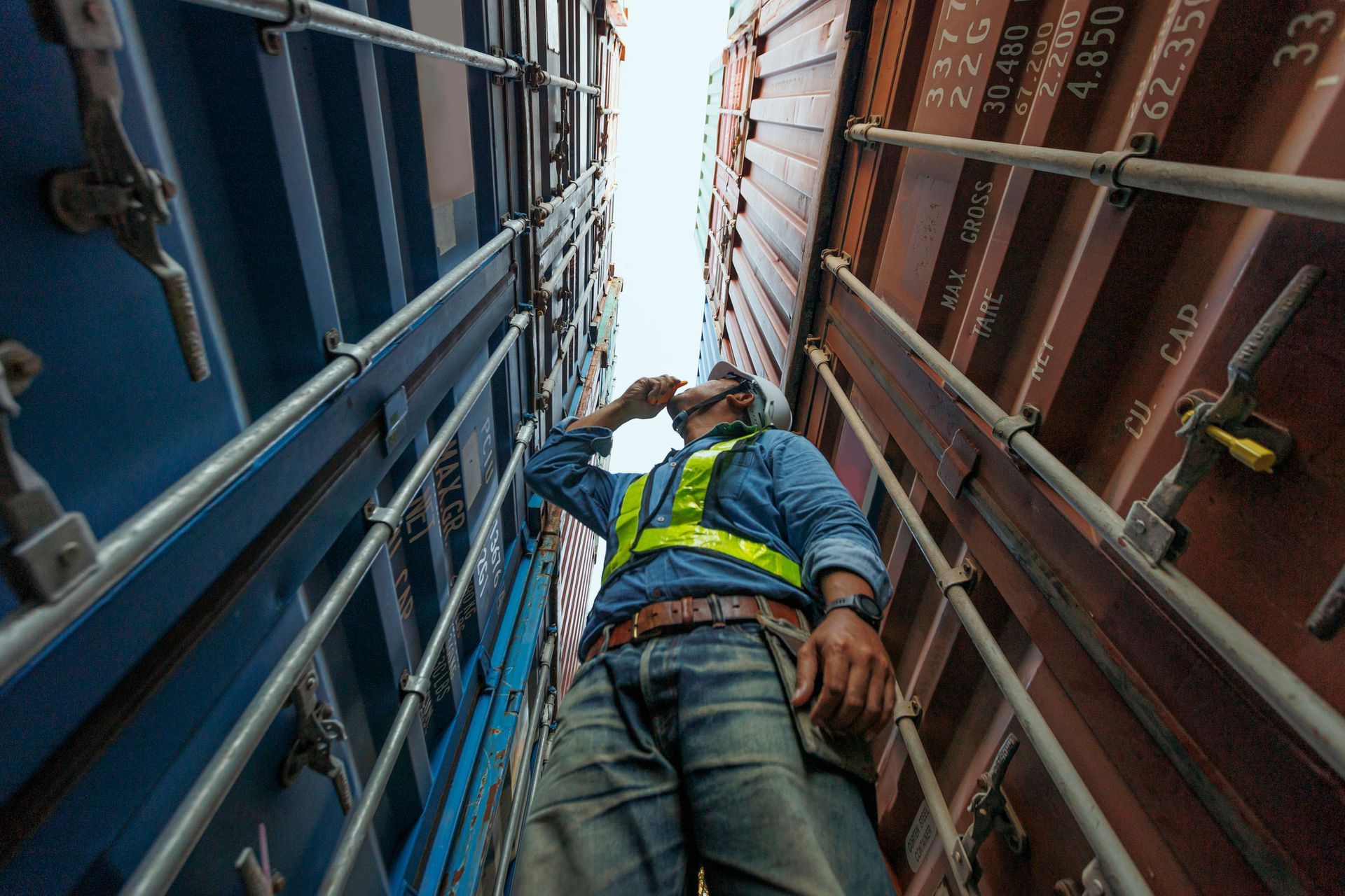 Worker in safety gear between containers, looking upward.