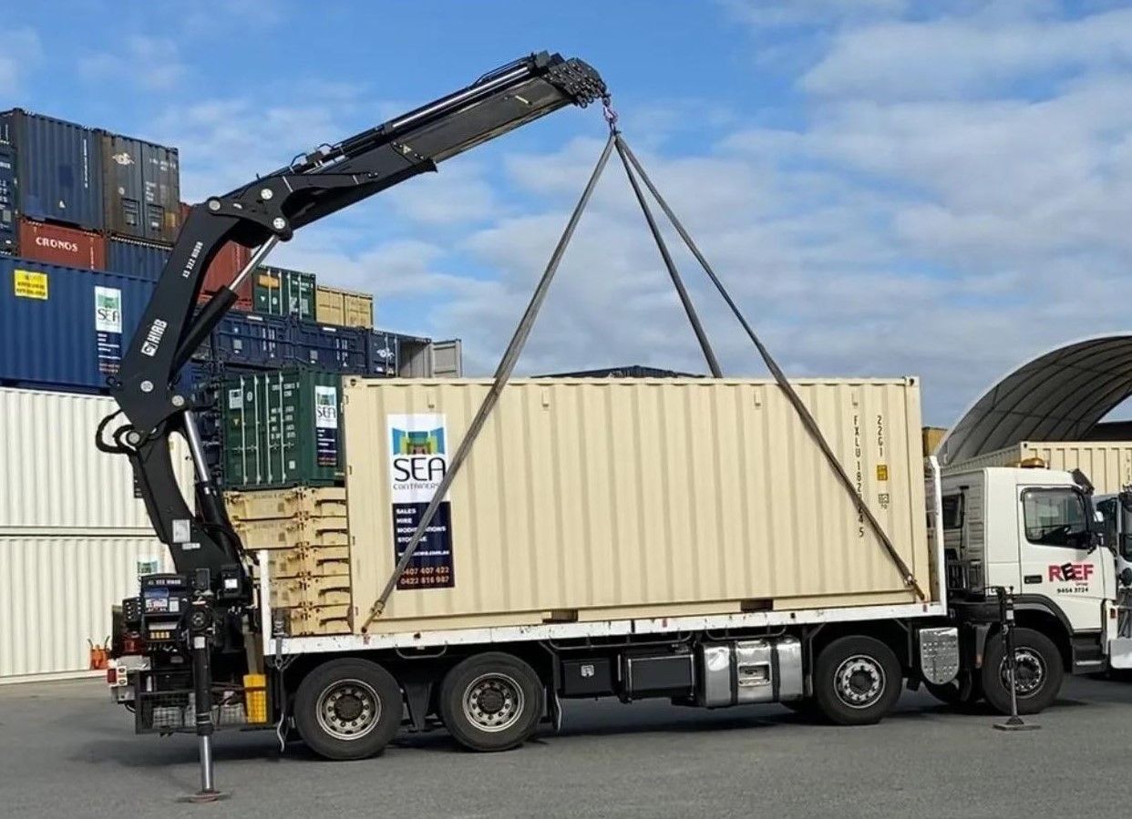 A crane lifting a tan cargo container onto a truck in an outdoor shipping yard.
