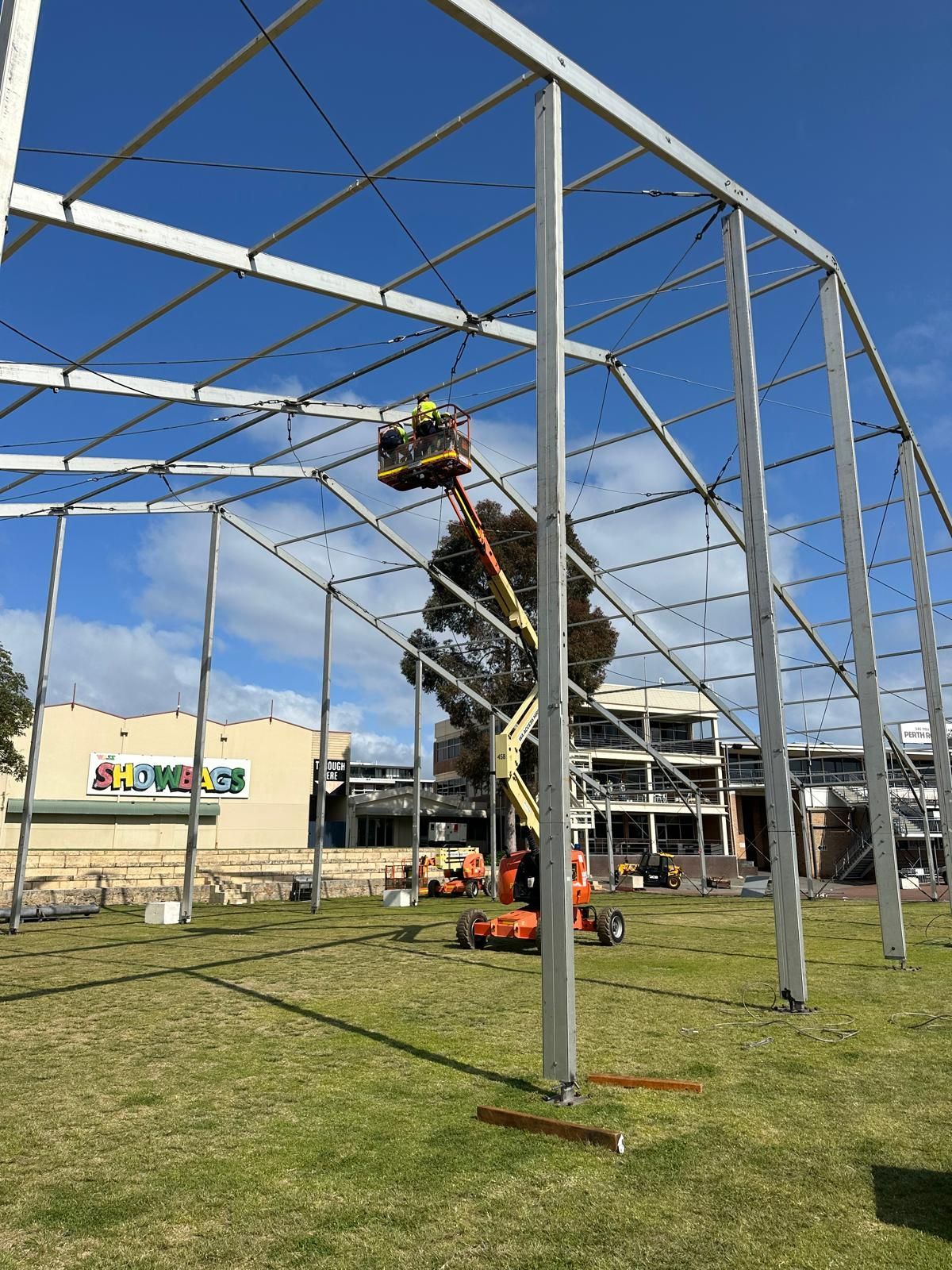 Worker in lift assembling a large metal frame outdoors on a green field under a blue sky.
