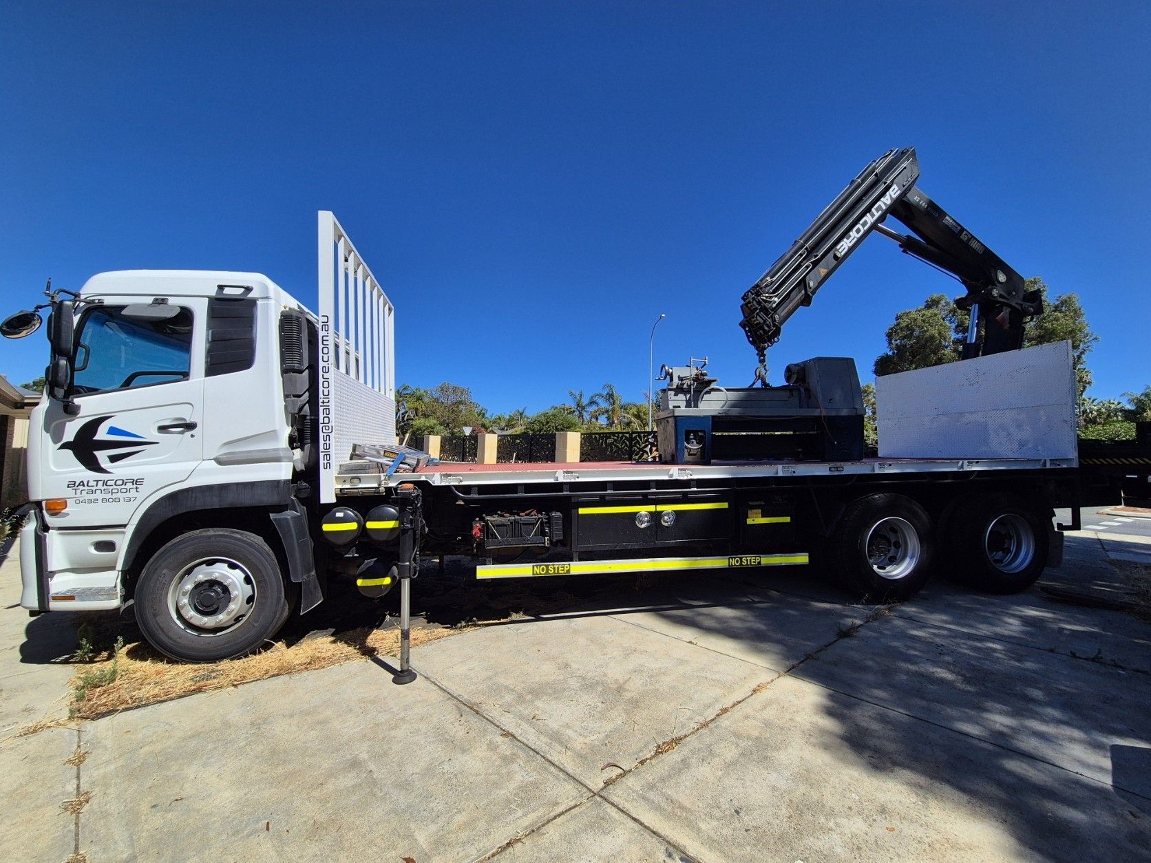 White flatbed truck with crane on a concrete driveway under a clear blue sky.
