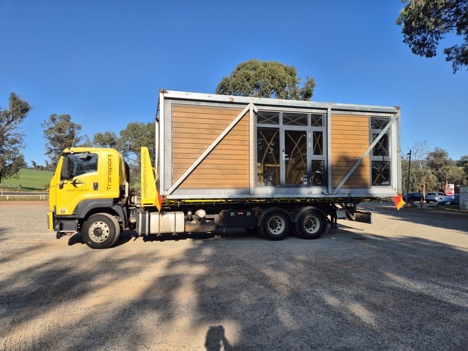 Yellow tow truck carrying a blue skid steer on its flatbed, parked on red dirt under a blue sky.