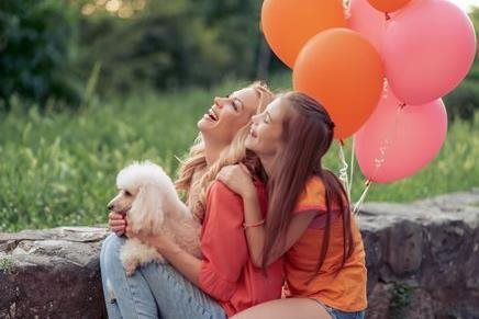 Two people sitting outdoors with a small white dog and pink balloons, smiling by a stone wall