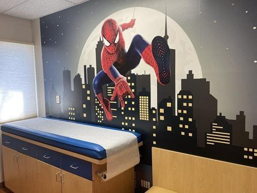 Spider-Man mural above a hospital exam table in a child-friendly room with city skyline art