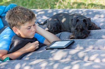 Boy lying on a gray cushion beside a black dog, looking at a tablet outdoors