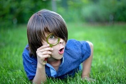 Child lying in grass, peering through a magnifying glass with a curious expression
