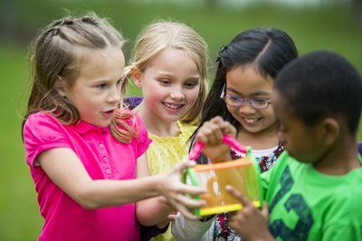 Children sharing a colorful outdoor activity on grass, smiling and reaching toward a toy structure
