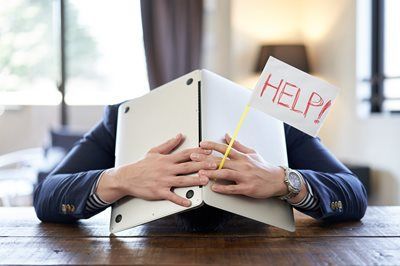 Person at desk hides face behind laptop while holding a “HELP!” sign