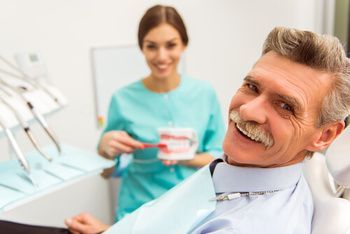 A Dentist in Teal Scrubs Holds a Model of Teeth While a Patient Smiles — Killara Dental in East Killara, NSW