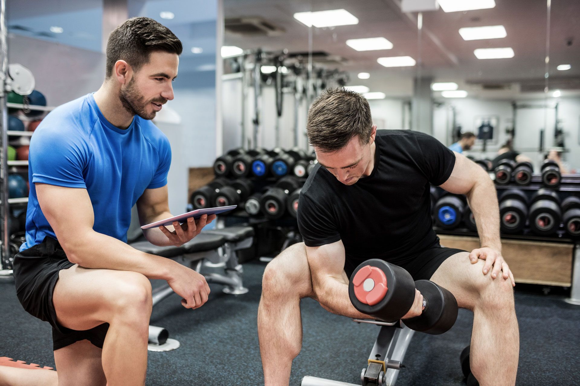 A man is lifting a dumbbell in a gym while another man looks at a tablet.