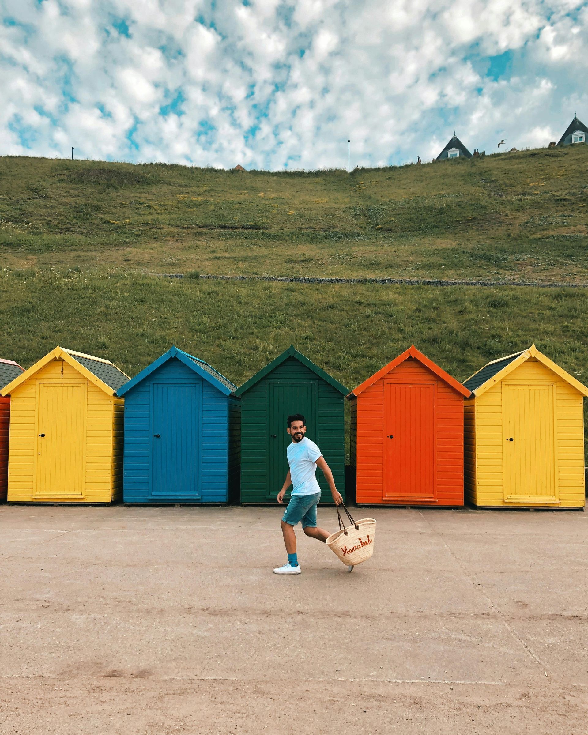 A happy man walks holding a basket, in front of a backdrop of multi-coloured painted sheds in Reading, England.