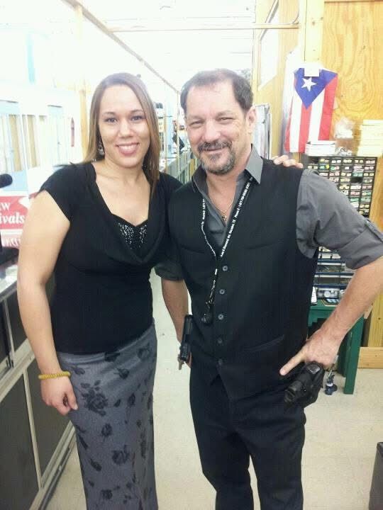 Woman in black top and gray skirt poses with man in vest. They smile inside a store.