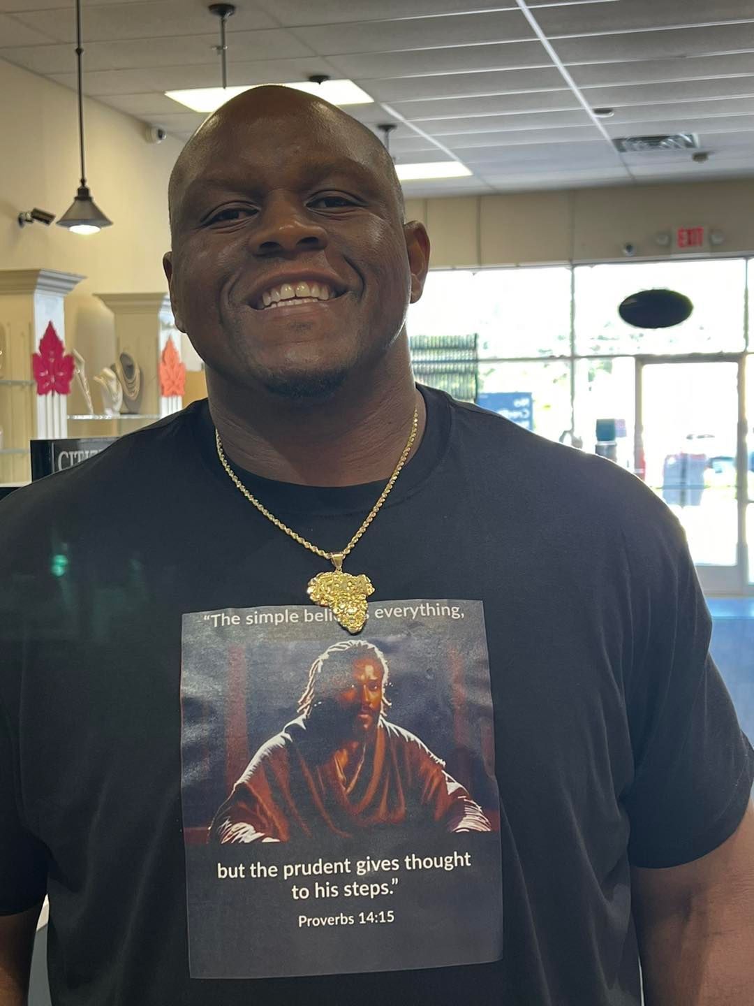 Man with a gold chain and t-shirt featuring a biblical image, smiling, in an indoor setting.