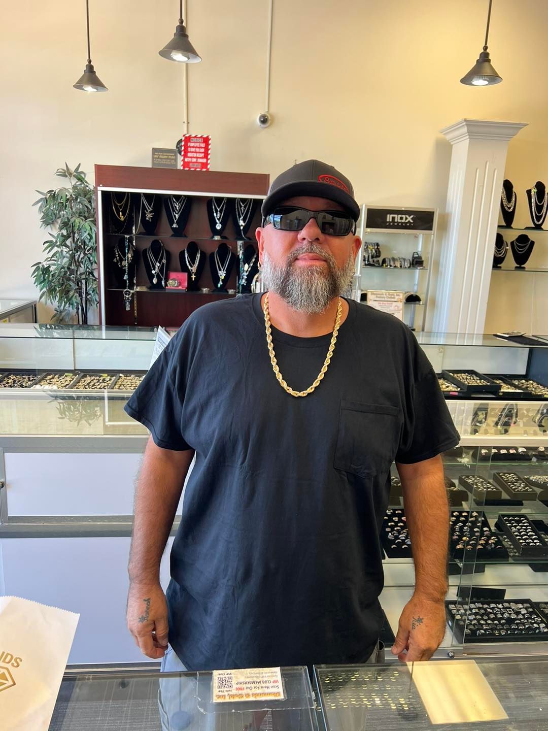 Man in jewelry store, wearing a chain, hat, and sunglasses, standing behind counter with displays of jewelry.