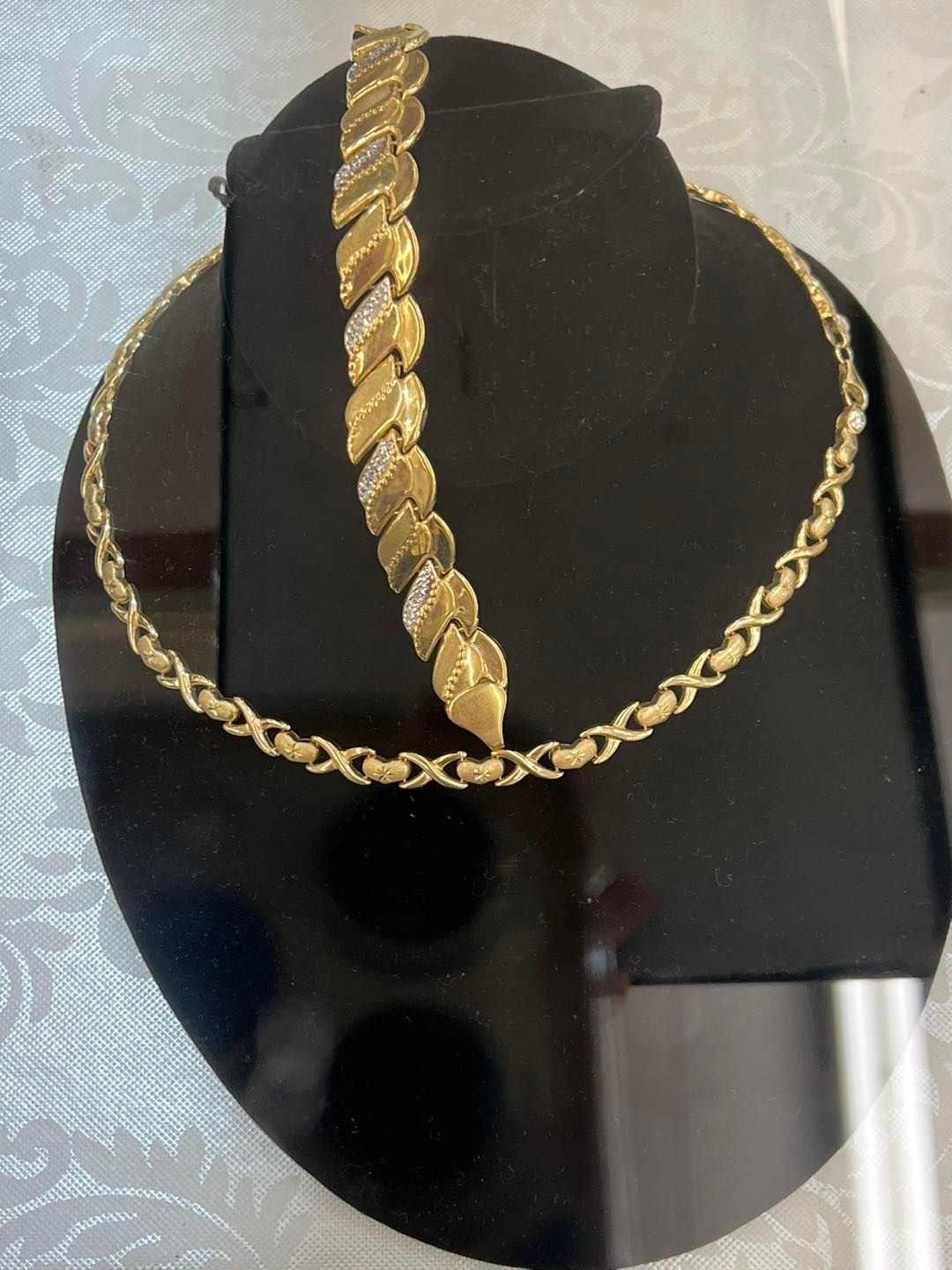 Gold necklace and bracelet on a black display stand; both pieces have a repeating pattern.